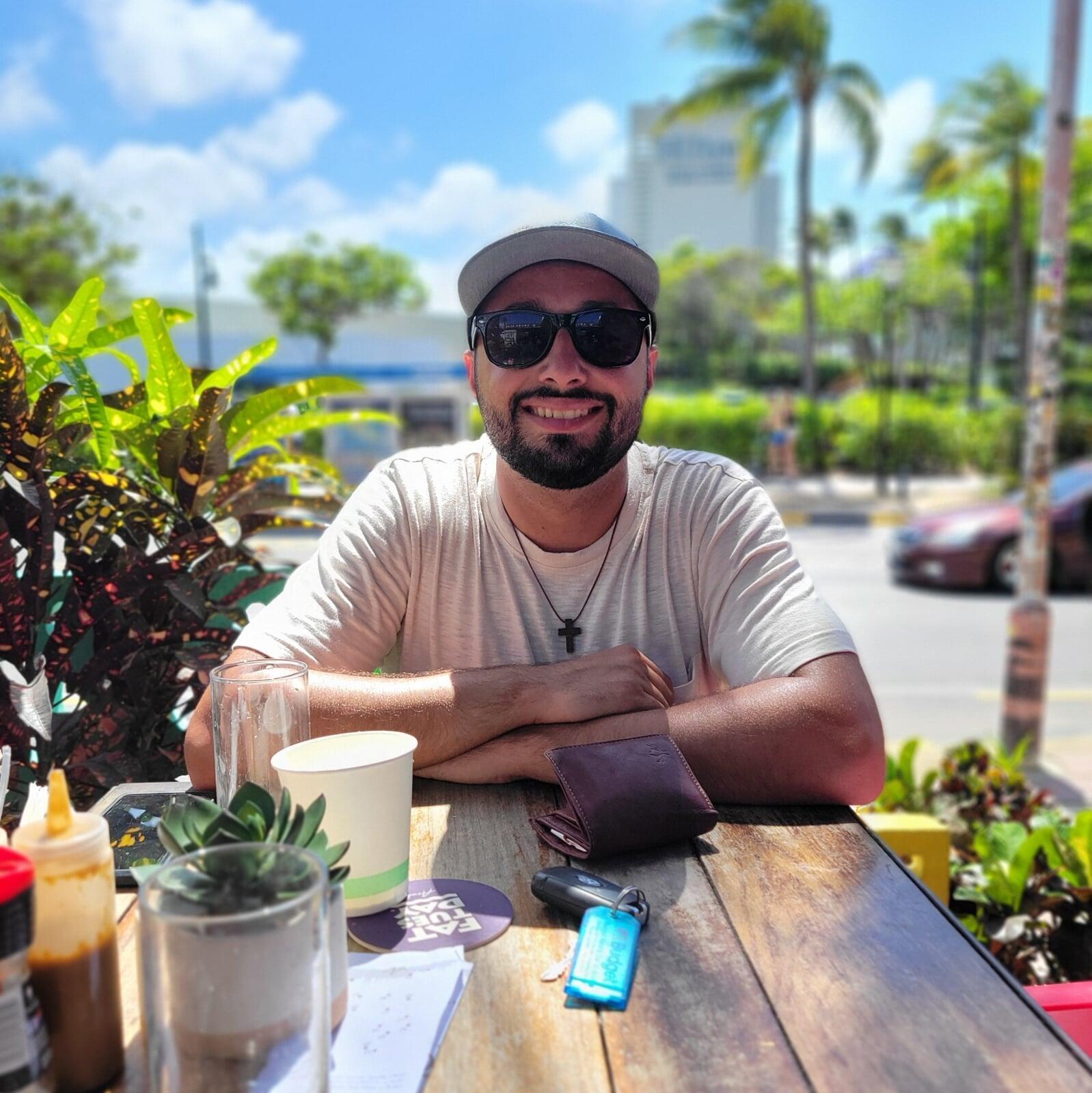 Man sitting at an outdoor café table smiling on a sunny day with palm trees in the background.